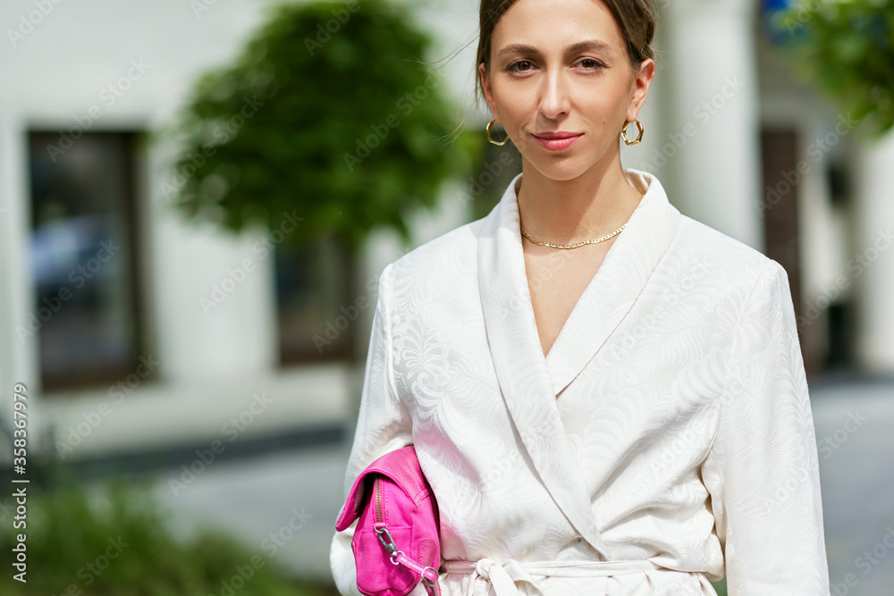 Outdoor portrait of stylish young woman in white suit and pink bag, walking down the street in the city on a sunny day. Pretty business female smiling and looking directly to the camera. Outdoor portrait of stylish young woman in white suit and pink bag, walking down the street in the city on a sunny day. Pretty business female smiling and looking directly to the camera.