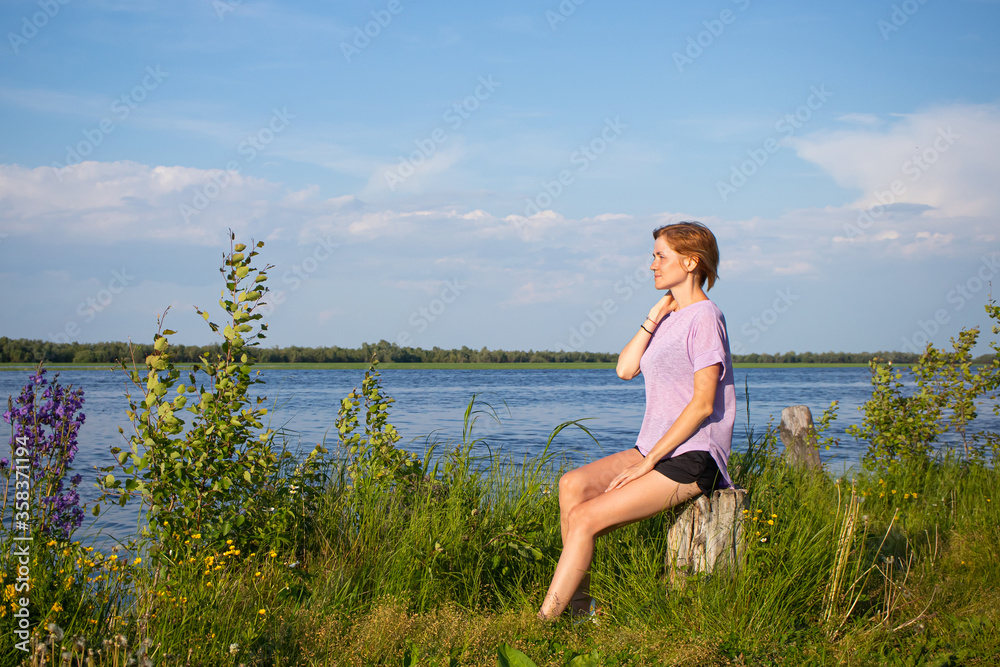 The concept of walks alone. Young girl with short hair in purple T-shirt and shorts sits on river bank. Summer sunny evening. Copyspace. Cozy cute rural landscape..