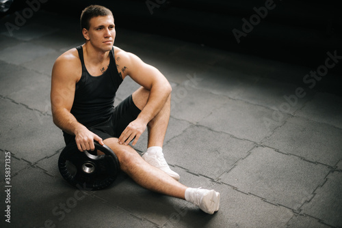 Muscular strong man with perfect beautiful body wearing sportswear sitting on the gym floor with weight from the barbell after heavy workout training in dark modern gym. Concept of healthy lifestyle.
