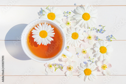 Chamomile tea in a white little cup on a light wooden surface. Around the cup are scattered flowers. The concept of natural vitamin drinks for health, immunity. Top view, flat lay..