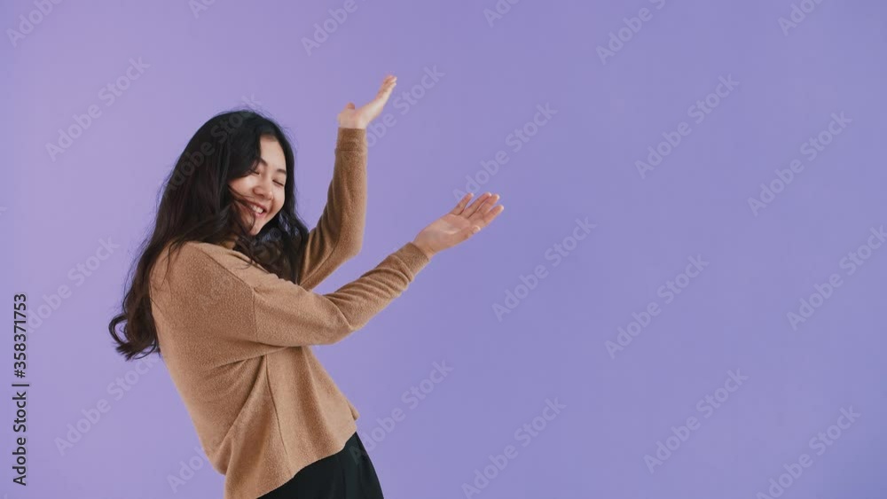 Asian woman in brown pullover. She smiling, pointing up by forefingers and showing thumbs up while posing against purple studio background. Close up Asian woman in brown pullover. She smiling, pointing up by forefingers and showing thumbs up while posing against purple studio background. Close up