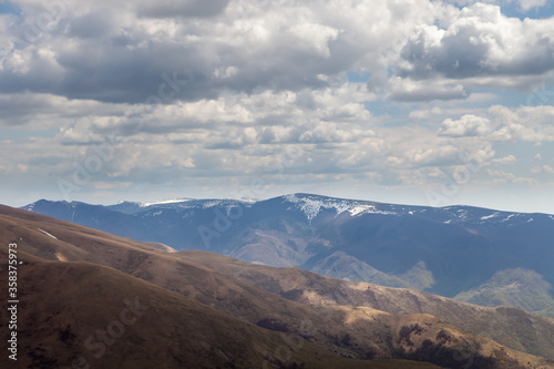 Rolling hills lighten by soft light, distant mountain peaks with remaining snow at spring and cloudy sky