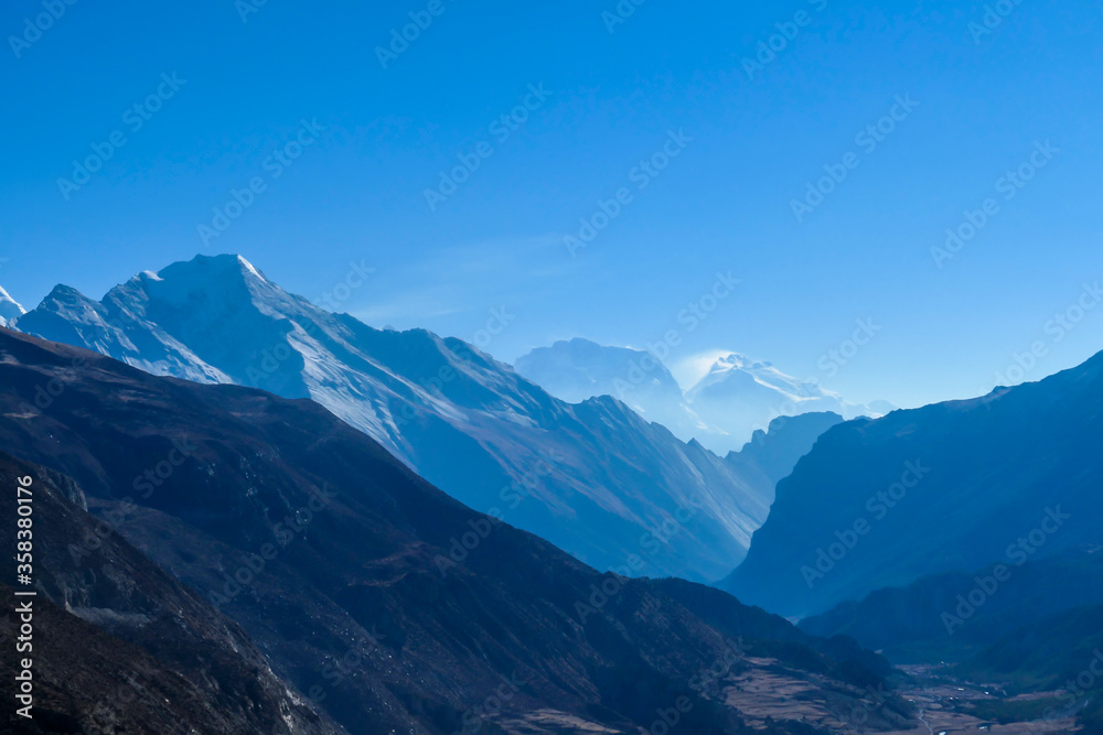 Naklejka View on high Himalayas along Annapurna Circuit Trek, Nepal. Harsh and barren landscape around. Clear and blue sky. High Himalayan ranges around. Snow capped mountains. Serenity and calmness