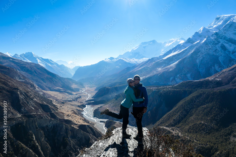 A couple standing at a mountain ledge, hugging and enjoying the view on Manang valley stretching in Himalayas, along Annapurna Circuit. Freedom. Love and passion. Snow capped Himalayas around.