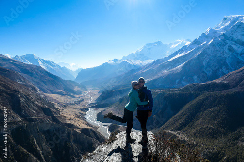 A couple standing at a mountain ledge, hugging and enjoying the view on Manang valley stretching in Himalayas, along Annapurna Circuit. Freedom. Love and passion. Snow capped Himalayas around.