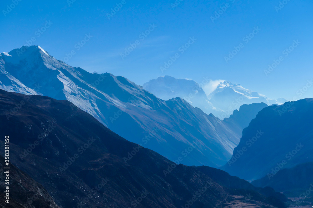 Naklejka View on high Himalayas along Annapurna Circuit Trek, Nepal. Harsh and barren landscape around. Clear and blue sky. High Himalayan ranges around. Snow capped mountains. Serenity and calmness