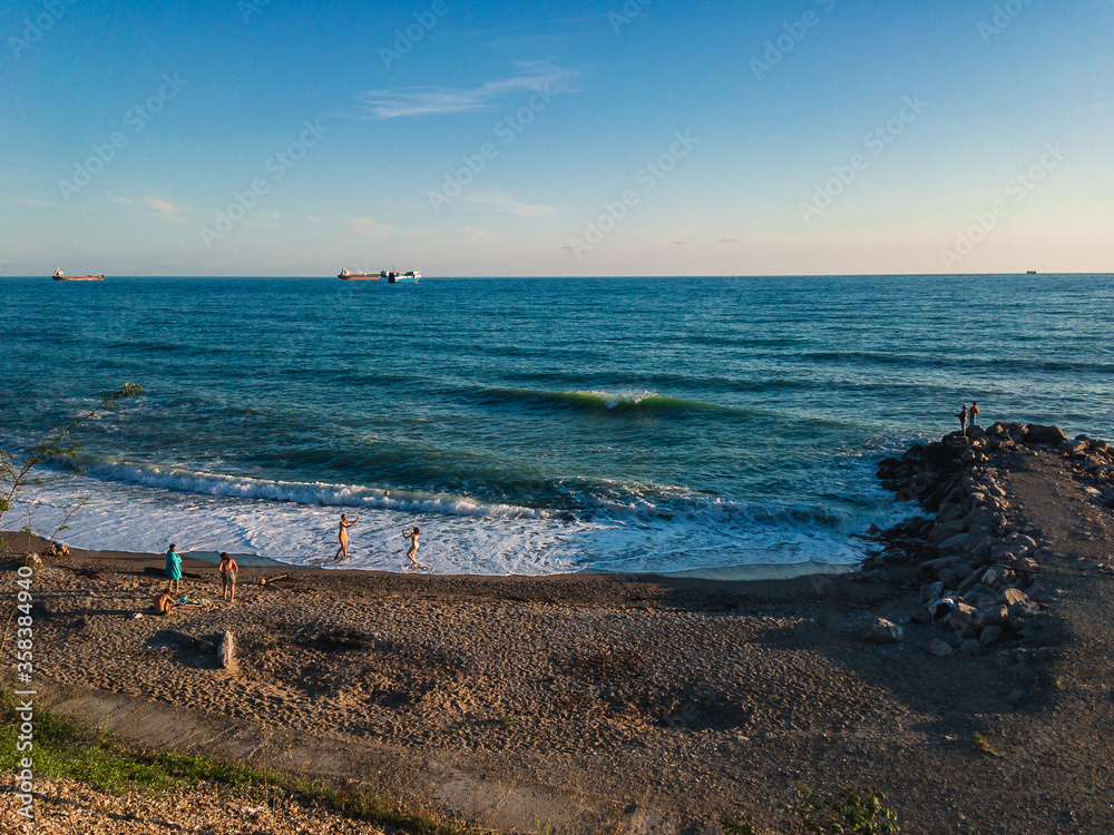 View of the Black Sea coast with people resting, lit by sunlight at sunset, with a breakwater and ships on the horizon against the blue sea on a sunny evening.