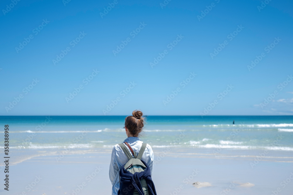Girl traveling with a backpack stands looking into the distance on a beach with white sand against a background of blue azure transparent water and a blue sky with a clear horizon line. Girl traveling with a backpack stands looking into the distance on a beach with white sand against a background of blue azure transparent water and a blue sky with a clear horizon line.