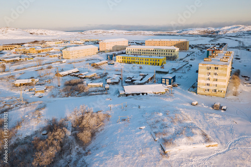 Aerial view to rural Teriberka cityscape in morning dawn light with panel buildings, wooden houses, garages, frosted trees, a lot of snow and rocky hills on coast of the Barents Sea in the background