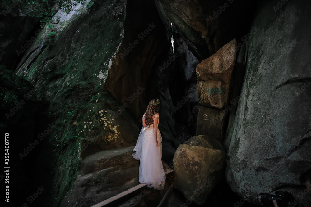 Young bride posing on a background of mountains. A smiling and beautiful bride in a white dress is standing against a background of rocks and trees. Wedding foto of a cute girl with a bouquet.