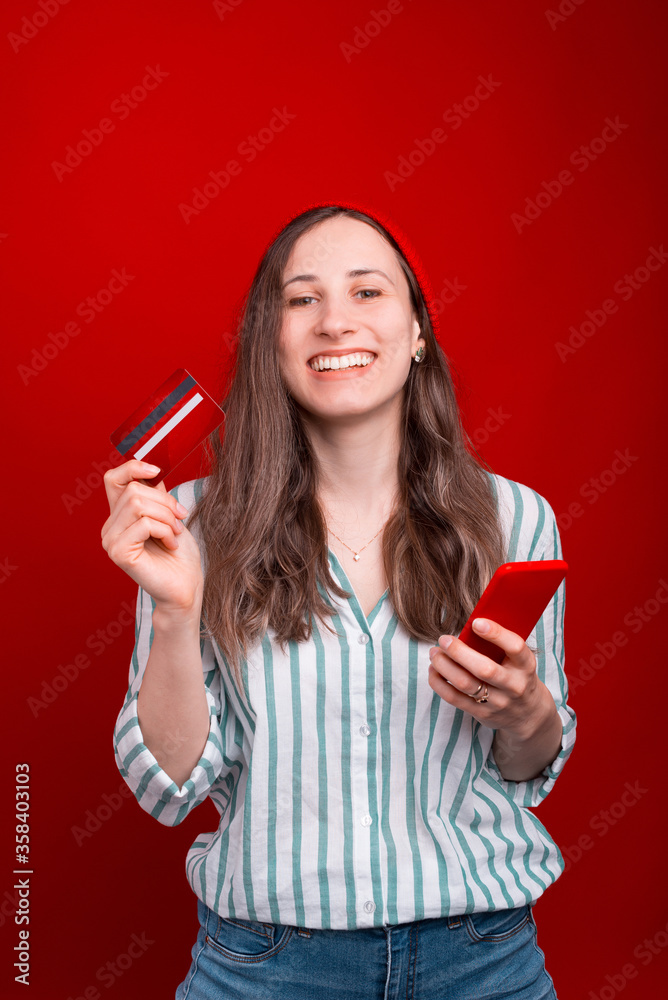 Smiling girl wearing white shirt is holding a phone and a card over red background Smiling girl wearing white shirt is holding a phone and a card over red background
