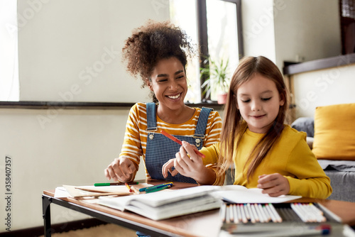 Caucasian little girl spending time with african american baby sitter. They are drawing, learning to write letters, sitting on the floor