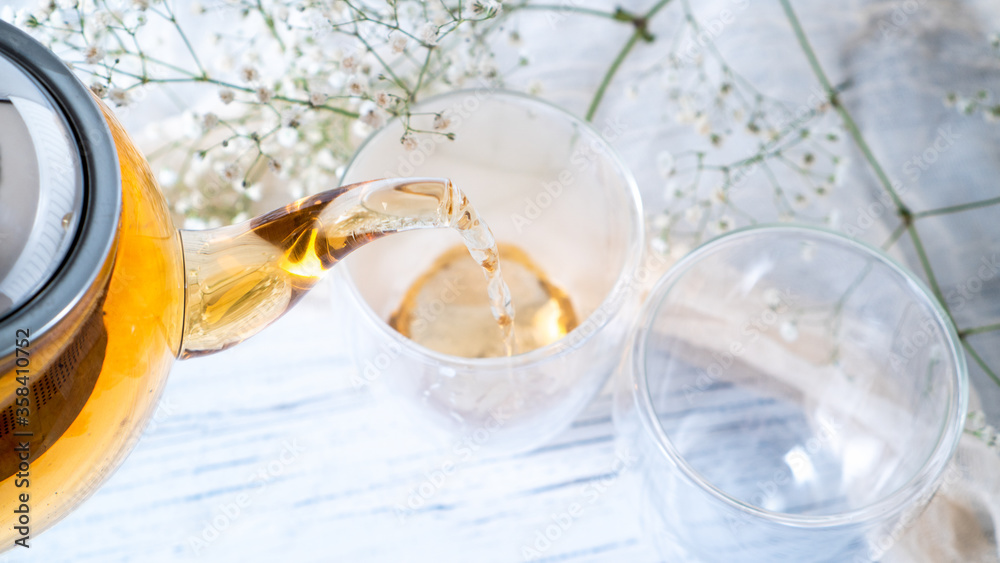 Pouring black tea from glass teapot into double wall cups. Golden tea. Empty mugs. Bright white wooden table.