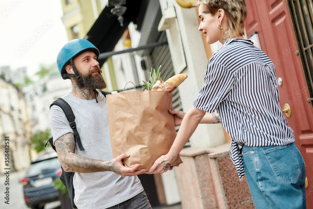 Bearded delivery man in helmet with backpack giving away grocery bag to a female customer, while delivering food. Courier, delivery service concept