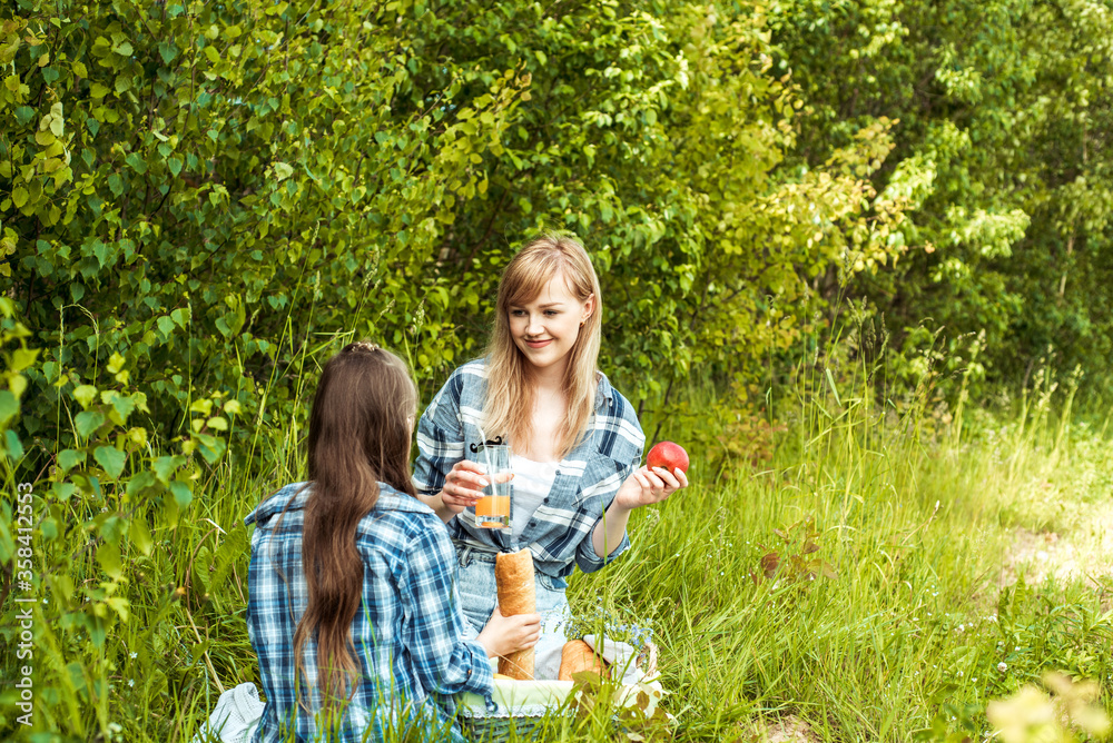 Obraz premium young mother and little daughters in bright sweaters on a picnic in nature indulge in apples.Picnic in the forest, green park, wicker basket, fruit, juice and white bread. Outdoors.