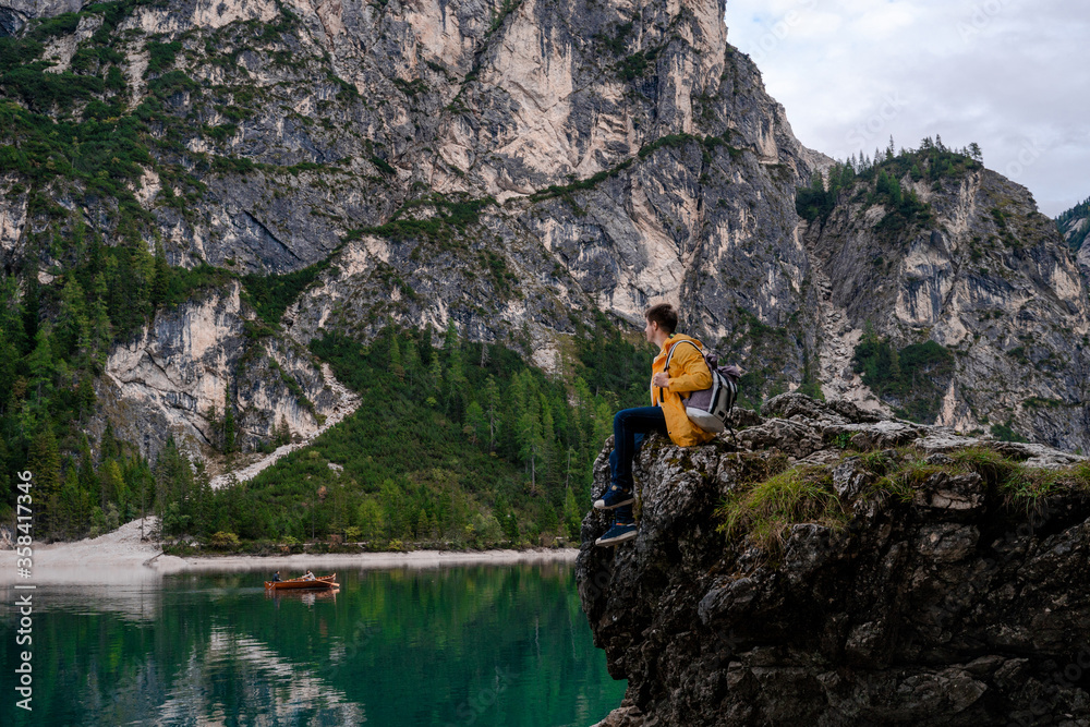 Obraz premium Brunet man in yellow raincoat sitting on the cliff and watching the boat floats on the lake Braies (Lago di Braies, Wildsee) on background of high mountain wall and cloudy sky in Dolomites Alps. Italy