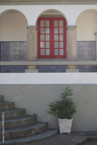 Janela vermelha numa entrada de uma casa antiga com parede em painel de azulejos, pilares em pedra, escada em pedra e um vaso com uma planta photo