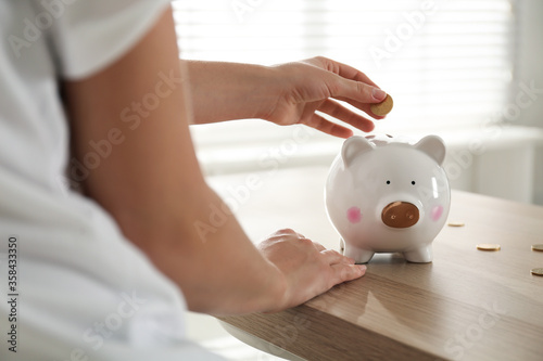 Woman putting money into piggy bank at wooden table indoors, closeup