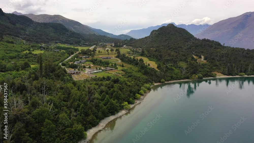 Aerial panning shot of trees at lakeshore near mountain range, drone flying forward over lake against sky - Queenstown, New Zealand