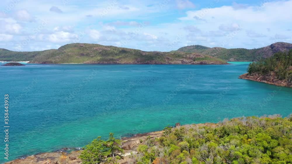 Aerial tilt down shot of mountains and sea against cloudy sky, drone ascending over green trees on rocky coastline during sunny day - Whitehaven Beach, Australia Aerial tilt down shot of mountains and sea against cloudy sky, drone ascending over green trees on rocky coastline during sunny day - Whitehaven Beach, Australia