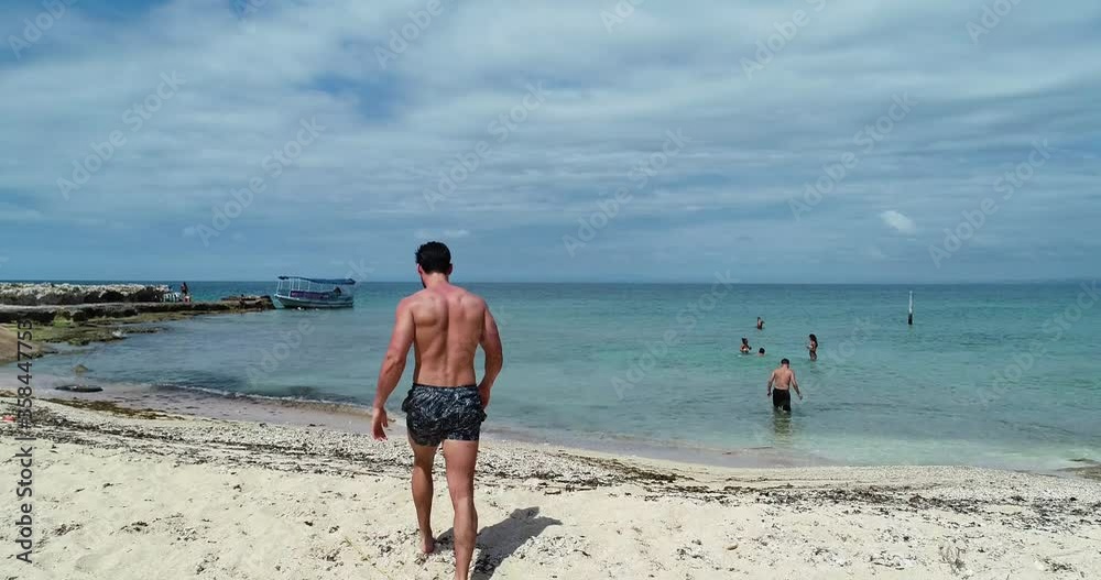 Young Caucasian strong athletic dark man wearing bathing suit shorts, shirtless and barefoot walks on sandy beach towards green ocean water, behind pan