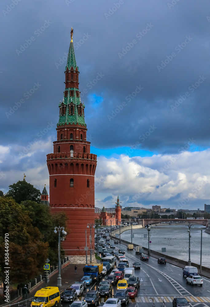 Fototapeta premium Red Kremlin wall and tower, traffic on Kremlevskaya Embankment by the Moskva River with a cloudy sky in Moscow, Russia