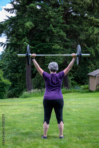 Middle aged caucasian woman with gray hair lifting a barbell with black plates, fitness outside on the lawn 
