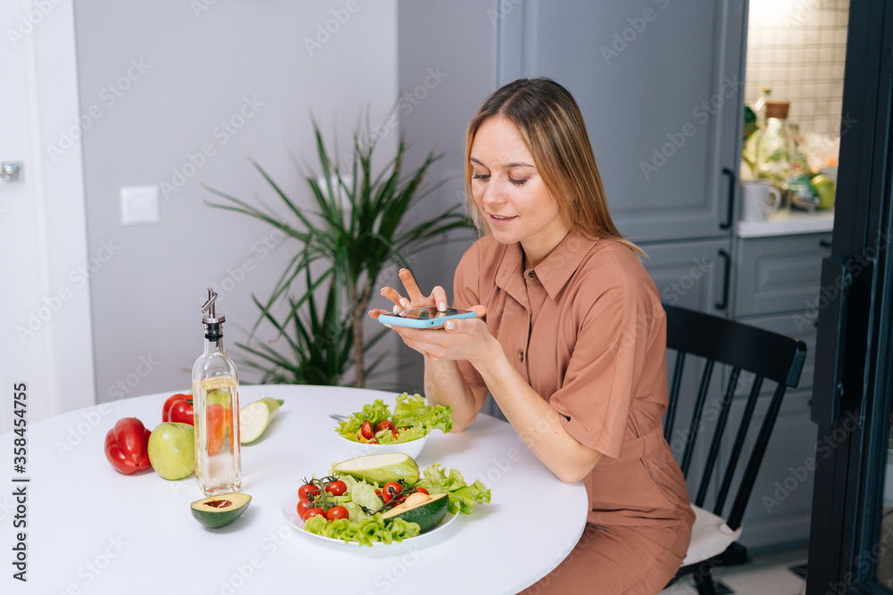 Happy young vegetarian woman uses smartphone for taking photos of vegetables lying on kitchen table. Concept of social networks. Female taking photos of vegetarian food on cell smart phone. Happy young vegetarian woman uses smartphone for taking photos of vegetables lying on kitchen table. Concept of social networks. Female taking photos of vegetarian food on cell smart phone.