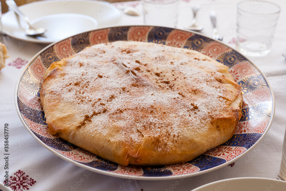 Food shot of a family size crispy delicious gourmet moroccan pastilla on an ornaments plate covered with icing sugar and cinnamon on a linens tablecloth with glasses, knife, fork and napkins. Food shot of a family size crispy delicious gourmet moroccan pastilla on an ornaments plate covered with icing sugar and cinnamon on a linens tablecloth with glasses, knife, fork and napkins.
