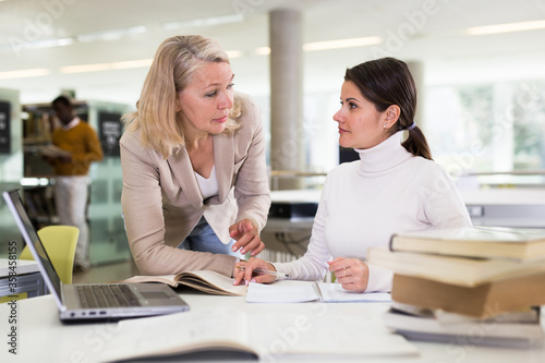 Friendly female tutor helping to diligent positive girl preparing for exam in library