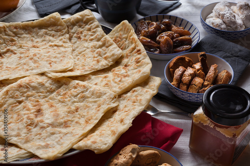 Rghayf Moroccan traditional squared semolina pancakes on a big plate on a white linens table background with dates, fekkas almond and raisins sweets and honey. Ramadan suhoor stills at home.