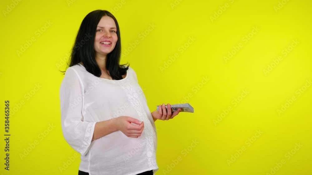 Rich young pregnant woman with black hair in a white blouse is standing with a bundle of money in her hand laughing and talking on a yellow background with copy space. Place for text or product Rich young pregnant woman with black hair in a white blouse is standing with a bundle of money in her hand laughing and talking on a yellow background with copy space. Place for text or product