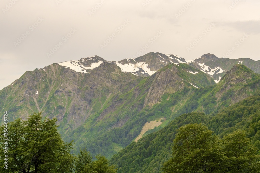 View of the snow-capped peaks of a large mountain range with spring-green slopes. Cloudy spring day.