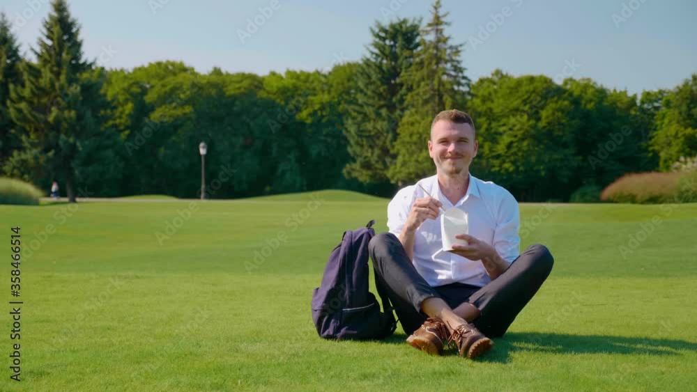 Man looks at the camera and smiles with a vegetarian lunch in an eco-friendly package. Guy is sitting in the park on the green grass, holding a paper package in his hand with a salad and wooden sticks Man looks at the camera and smiles with a vegetarian lunch in an eco-friendly package. Guy is sitting in the park on the green grass, holding a paper package in his hand with a salad and wooden sticks