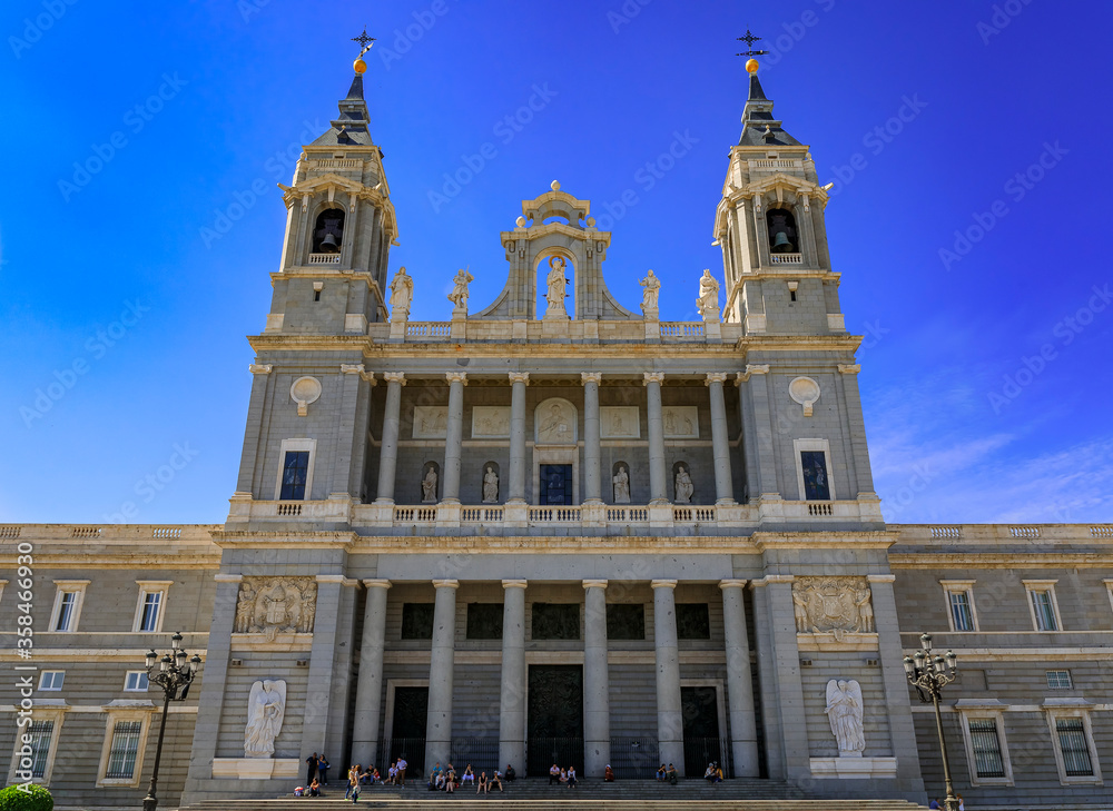Main facade of the Cathedral of Our Lady of La Almudena by the Royal Palace, people sitting on steps in Madrid, Spain