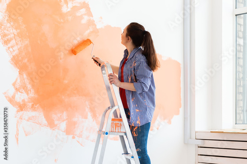 Beautiful female painting the wall with paint roller. Portrait of a young beautiful woman painting wall in her new apartment. Redecoration and renovation concept.
