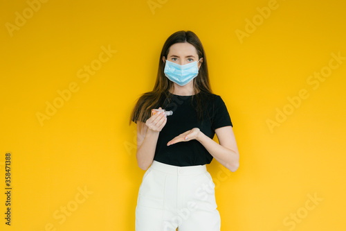 Fashion portrait of young woman wearing a face mask, looking at camera, use antiseptic, isolated on yellow background. Flu epidemic, dust allergy, protection against virus. Corona virus. COVID-19.