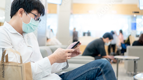Male Asian patient wearing surgical face mask using smartphone at waiting area in hospital or medical centre. Medical exam or body check up. Coronavirus pandemic prevention. Health care concept