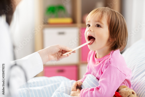 healthcare, medicine and people concept - doctor checking sick little girl's throat with tongue depressor in bed at home photo