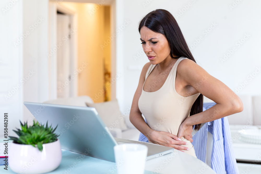 Businesswoman Holding Her Back While Working On Laptop At Home Office Desk. young woman, stretching, sitting feeling her back tired after working at laptop, small home office interior. Businesswoman Holding Her Back While Working On Laptop At Home Office Desk. young woman, stretching, sitting feeling her back tired after working at laptop, small home office interior.