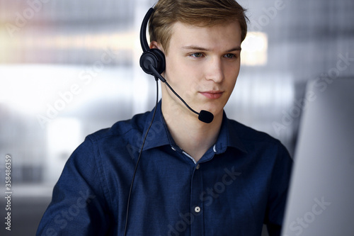 Young blond businessman using headset and computer in a darkened office, glare of light on the background. Startup business means working hard