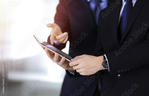 Business people use a tablet computer for discussion of their new project, standing in sunny modern office. Unknown businessman or male entrepreneur with a colleague at workplace