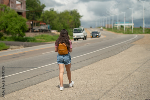 Girl tourists with backpacks hitchhiking on road. Hitchhiking tourism concept. Travel hitchhiker girl walking on road. Girl in the pink t-shirt and denim shorts walking near the road.