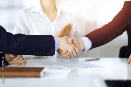 Business people shaking hands at meeting or negotiation, close-up. Group of unknown businessmen, and a woman on the background in a sunny modern office. Teamwork, partnership and handshake concept