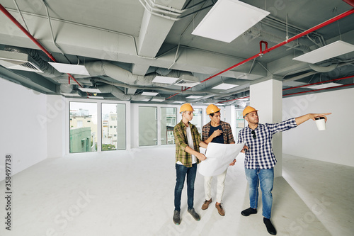 Three professional construction engineers wearing hardhats standing together inside new building planning further work, copy space