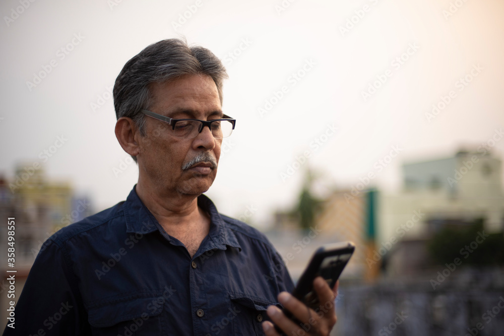 An old / aged Indian Bengali man in blue shirt is watching his cellphone while standing on a rooftop under the open sky. Indian lifestyle and seniors An old / aged Indian Bengali man in blue shirt is watching his cellphone while standing on a rooftop under the open sky. Indian lifestyle and seniors