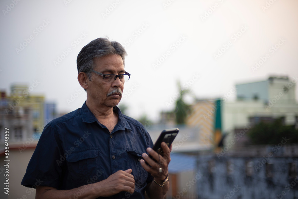 Fototapeta premium An old / aged Indian Bengali man in blue shirt is watching his cellphone while standing on a rooftop under the open sky. Indian lifestyle and seniors