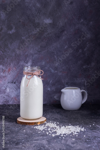 Vegan rice milk in a glass bottle and rice in a white plate on a wooden stand with a molon on a gray background photo