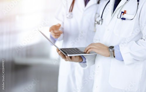 Two unknown doctors with stethoscopes discuss medical exam resoults, whille standing at sunny hospital office. Physicians use a laptop for checking up medication names records. Perfect medical service
