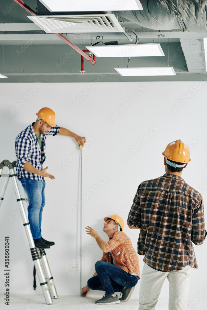 Vertical shot of professional construction worker standing on stepladder holding tape rule measuring wall length, his colleagues helping him, copy space Vertical shot of professional construction worker standing on stepladder holding tape rule measuring wall length, his colleagues helping him, copy space